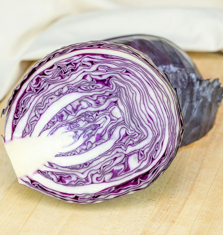 Chou Integro coupé en deux sur une surface de bois clair. L’intérieur montre des feuilles blanches et pourpres repliées en couches serrées autour d’un cœur blanc court. Une pomme entière violette apparaît derrière la coupe. Un tissu clair occupe le fond de l’image et souligne les contrastes entre les nervures pâles et les plis colorés.