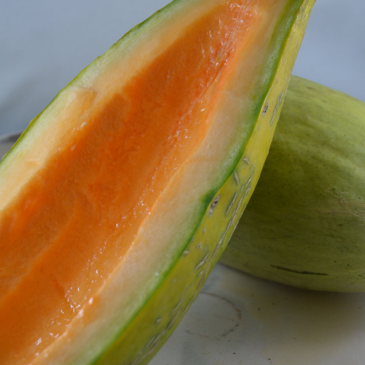 Melon Banana. Melon allongé coupé dans la longueur, à chair orange. Le cadrage permet de voir les formes, les couleurs et les détails du sujet représenté. Les contours se détachent du fond de l’image, avec des différences de teinte et de texture visibles sur les surfaces. Cette vue accompagne la présentation du produit dans la boutique.