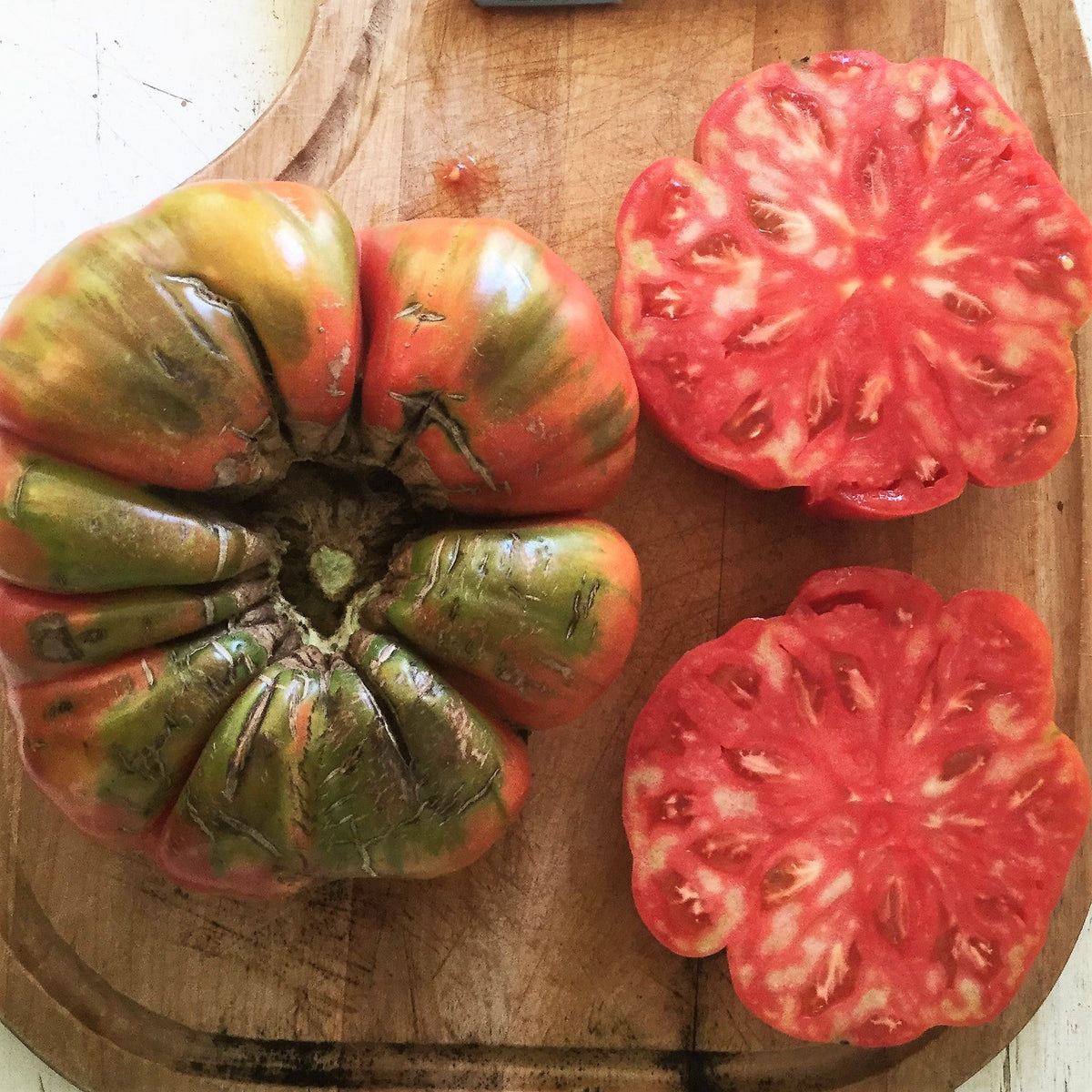 Tomate Beefsteak Adelin Morin. Grosses tomates côtelées, entières et coupées sur une chair rouge. Le cadrage permet de voir les formes, les couleurs et les détails du sujet représenté. Les contours se détachent du fond de l’image, avec des différences de teinte et de texture visibles sur les surfaces. Cette vue accompagne la présentation du produit dans la boutique.