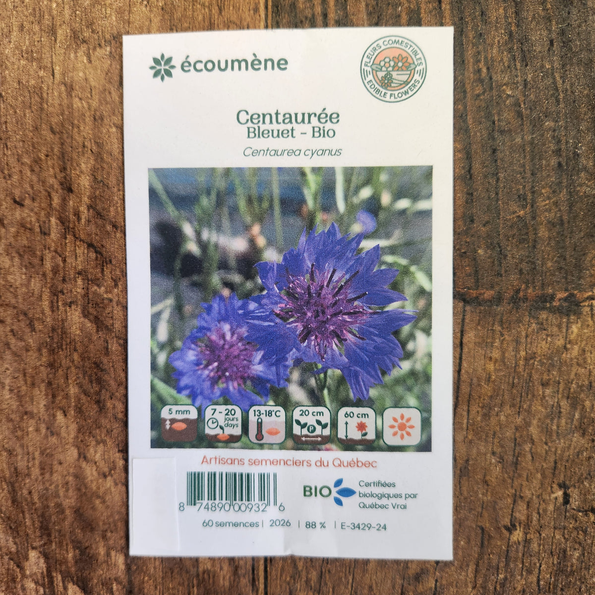 Sachet de semences Centaurée bleuet. Emballage blanc photographié de face sur un fond de bois brun. Une image du végétal occupe la partie centrale, sous le nom imprimé. De petits pictogrammes et un code-barres apparaissent au bas du sachet. La marque Écoumène figure dans la partie supérieure de cette présentation.