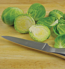 Choux de Bruxelles Nautic sur une planche en bois clair. Deux bourgeons coupés montrent leurs feuilles pâles serrées au centre, tandis que des choux verts entiers se trouvent derrière. Quelques feuilles détachées sont disposées à droite. Un couteau de cuisine traverse le premier plan, avec sa lame pointant vers la gauche de l’image.