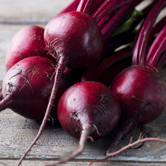 Des betteraves rouge foncé reposent sur des planches grises. Leurs racines fines s’allongent vers le bas et leurs pétioles pourpres se regroupent dans le haut droit. Certains légumes sont ronds et d’autres plus allongés. Des reflets éclairent la peau, tandis que les lignes du bois et les ombres restent visibles entre les racines du premier plan.