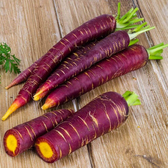 Des carottes violettes sont disposées en diagonale sur des planches brunes. Deux portions coupées montrent un centre jaune orangé entouré d’un bord pourpre. De fines lignes claires et de petites racines marquent les surfaces longues. Les bases vertes dépassent dans le haut droit, tandis que les pointes effilées et les faces coupées occupent le premier plan.