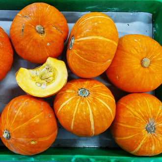 Des courges orange arrondies sont disposées dans une caisse verte. Des lignes jaunes ou crème traversent leur peau et des attaches gris-vert marquent les extrémités. Une portion coupée montre une chair jaune pâle et une cavité contenant des graines. Le fond du contenant est visible entre les fruits, qui se chevauchent dans cette présentation rapprochée.