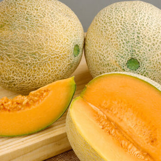 Deux melons entiers à peau beige réticulée accompagnent une moitié de melon coupée. La face ouverte montre une chair orange autour d’un creux contenant des graines claires. Les fruits sont disposés sur un support clair, avec les surfaces entières derrière la portion coupée. Les motifs de l’écorce et la couleur de la chair créent le contraste principal de l’image.