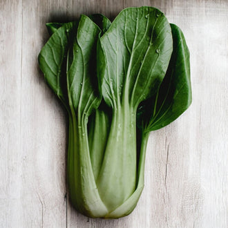 Un pak choi est posé sur des planches gris clair. Les pétioles épais vert très pâle se regroupent dans une base élargie et montent vers de grandes feuilles vert foncé. Les limbes se superposent dans le haut, avec des nervures plus claires. Le support reste visible autour du plant isolé, accompagné d’une ombre discrète sur le bois.