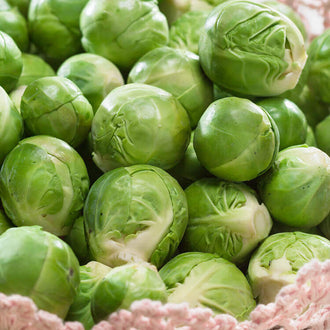 Un amas de petits choux de Bruxelles verts remplit le cadrage. Les feuilles serrées de chaque chou présentent des plis et des nervures pâles. Plusieurs extrémités coupées plus claires sont visibles dans le groupe. Une matière rose très pâle borde le bas de l’image, tandis que les légumes superposés forment des zones d’ombre entre leurs contours arrondis.