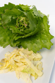Une pomme de chicorée aux grandes feuilles vertes et ondulées occupe le haut de l’image sur un fond blanc. Au premier plan, des lanières jaune pâle sont ponctuées de petites taches roses et rouges. Des nuances rosées bordent aussi certaines feuilles du cœur, tandis que les nervures restent discrètes sur les parties vertes.
