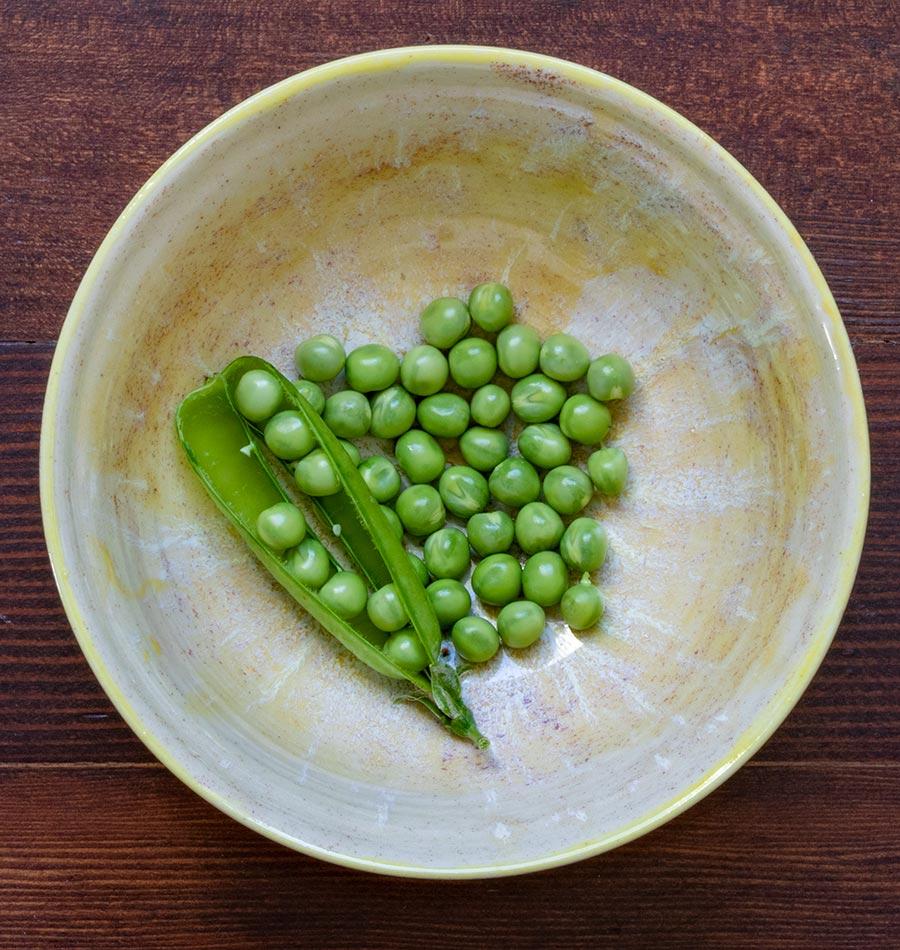 Un bol rond jaune pâle repose sur une table de bois brun. Il contient des petits pois verts écossés et une gousse ouverte, placée sur le côté gauche. Les grains sont regroupés au centre du bol, dont la surface présente des nuances crème et dorées. Cette vue de dessus illustre la récolte du pois Alaska.
