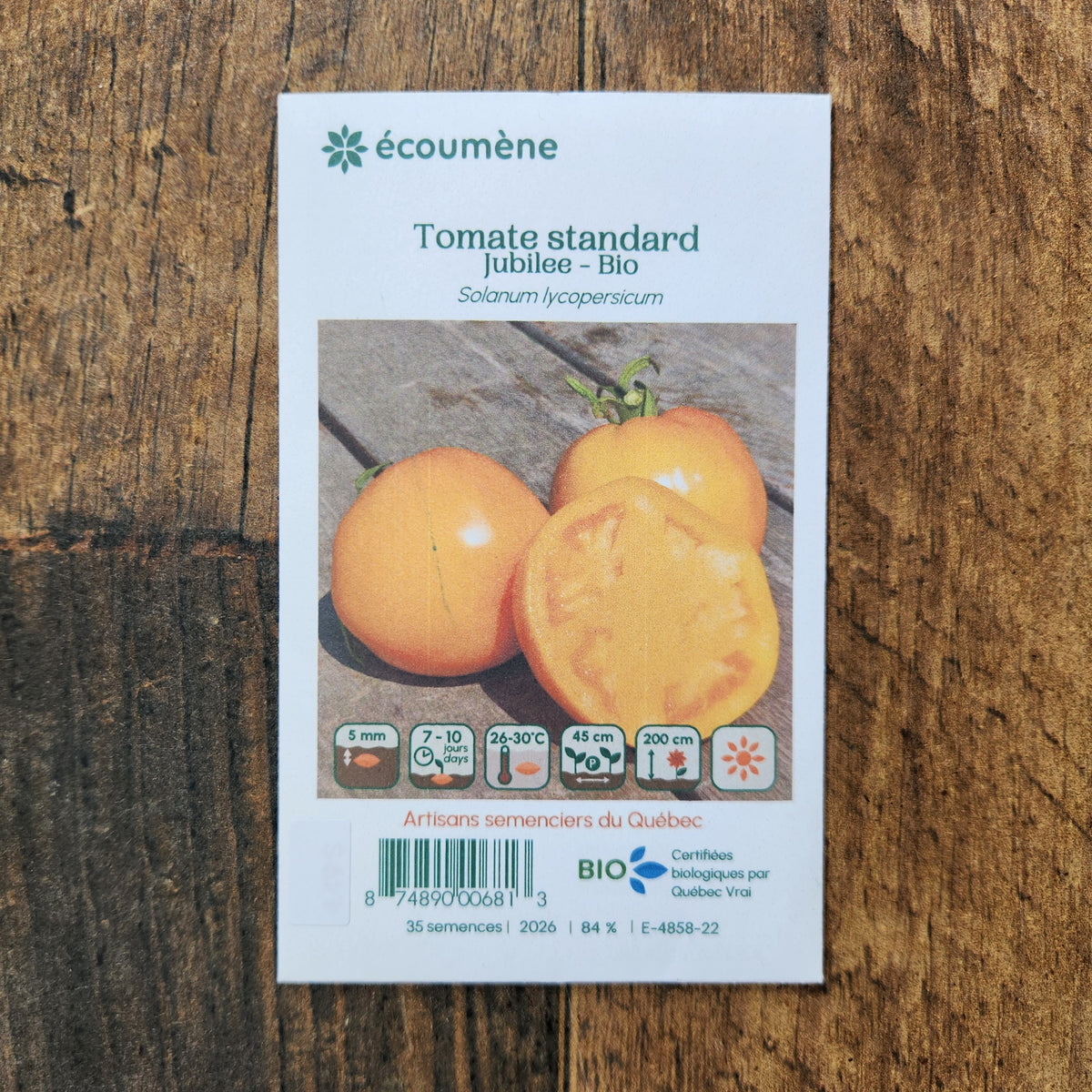 Sachet de semences Tomate Jubilee. Emballage blanc photographié de face sur un fond de bois brun. Une image du végétal occupe la partie centrale, sous le nom imprimé. De petits pictogrammes et un code-barres apparaissent au bas du sachet. La marque Écoumène figure dans la partie supérieure de cette présentation.