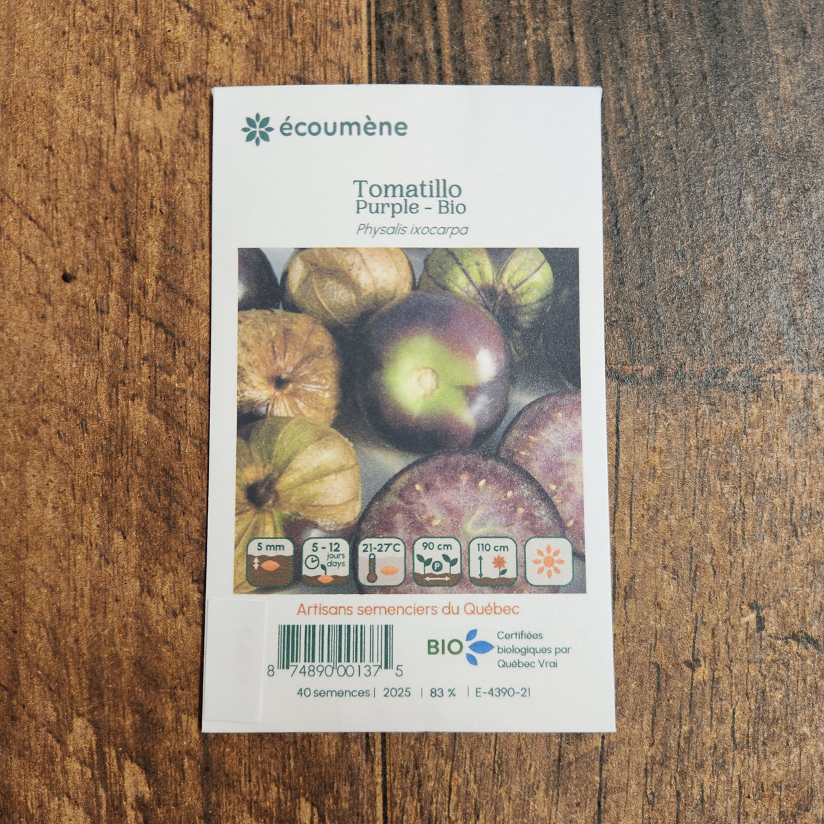 Sachet de semences Tomatillo Purple. Emballage blanc photographié de face sur un fond de bois brun. Une image du végétal occupe la partie centrale, sous le nom imprimé. De petits pictogrammes et un code-barres apparaissent au bas du sachet. La marque Écoumène figure dans la partie supérieure de cette présentation.