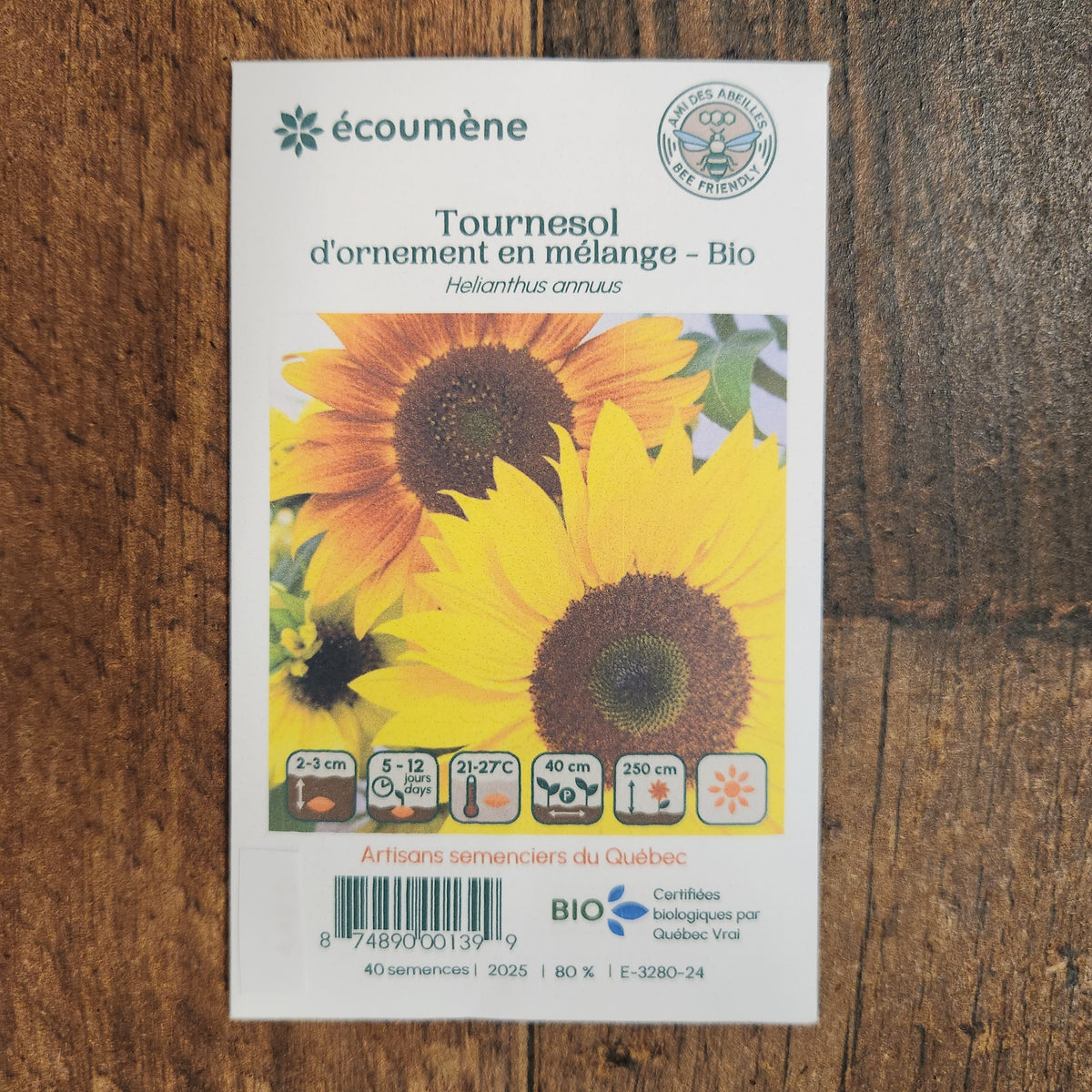 Sachet de semences Mélange de tournesol. Emballage blanc photographié de face sur un fond de bois brun. Une image du végétal occupe la partie centrale, sous le nom imprimé. De petits pictogrammes et un code-barres apparaissent au bas du sachet. La marque Écoumène figure dans la partie supérieure de cette présentation.