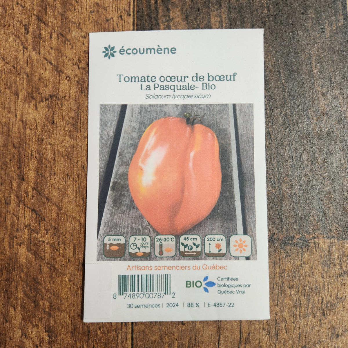 Sachet de semences Tomate Cœur de Bœuf La Pasquale. Emballage blanc photographié de face sur un fond de bois brun. Une image du végétal occupe la partie centrale, sous le nom imprimé. De petits pictogrammes et un code-barres apparaissent au bas du sachet. La marque Écoumène figure dans la partie supérieure de cette présentation.
