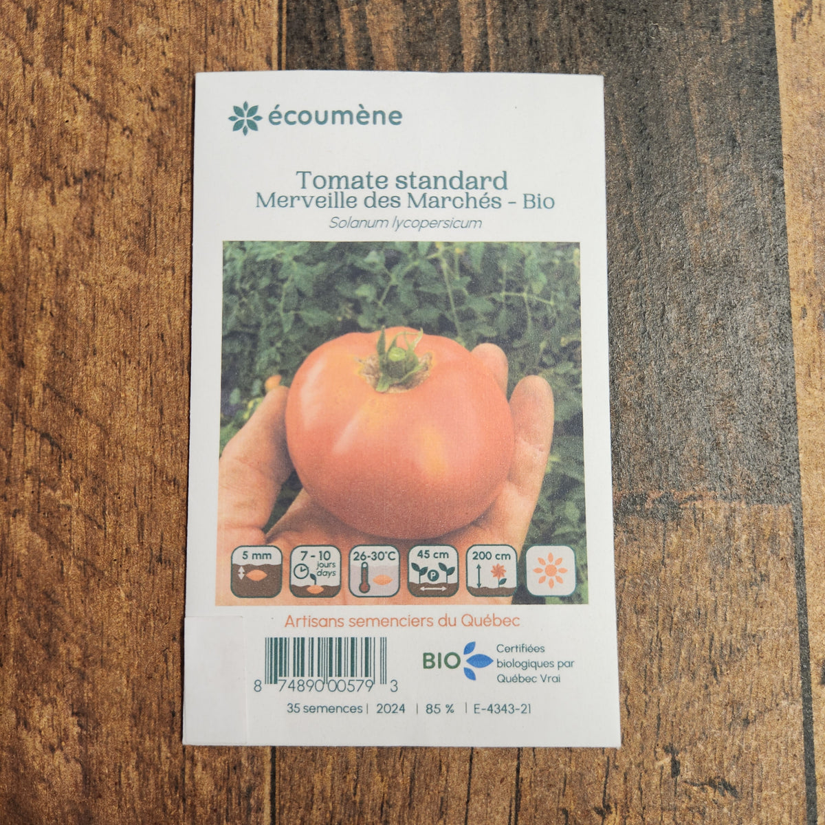 Sachet de semences Tomate Merveille des Marchés. Emballage blanc photographié de face sur un fond de bois brun. Une image du végétal occupe la partie centrale, sous le nom imprimé. De petits pictogrammes et un code-barres apparaissent au bas du sachet. La marque Écoumène figure dans la partie supérieure de cette présentation.