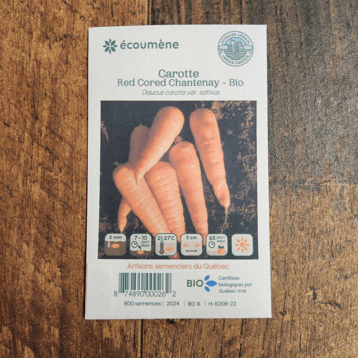 Sachet de semences Carotte Red Cored Chantenay. Emballage blanc photographié de face sur un fond de bois brun. Une image du végétal occupe la partie centrale, sous le nom imprimé. De petits pictogrammes et un code-barres apparaissent au bas du sachet. La marque Écoumène figure dans la partie supérieure de cette présentation.
