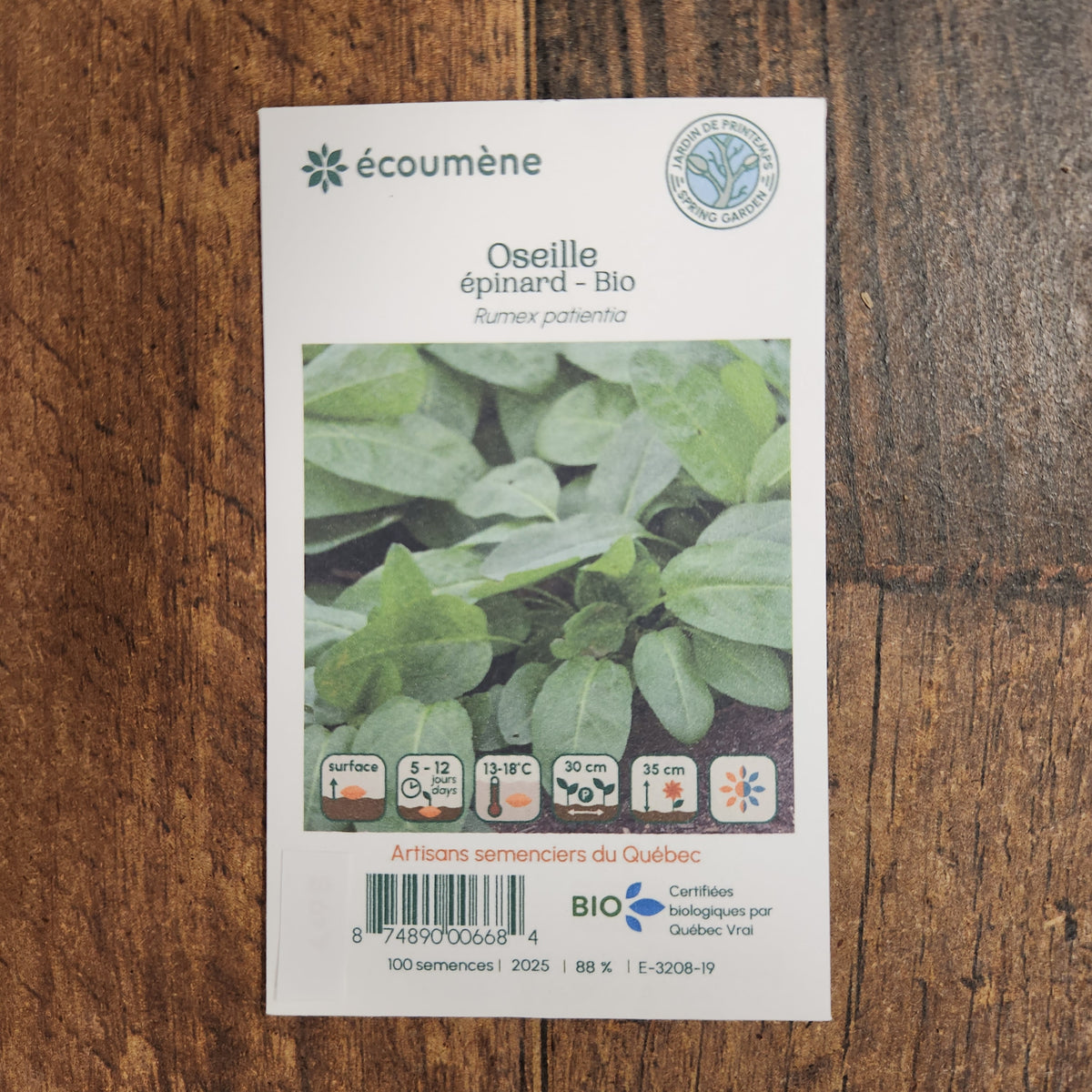 Sachet de semences Oseille Épinard. Emballage blanc photographié de face sur un fond de bois brun. Une image du végétal occupe la partie centrale, sous le nom imprimé. De petits pictogrammes et un code-barres apparaissent au bas du sachet. La marque Écoumène figure dans la partie supérieure de cette présentation.