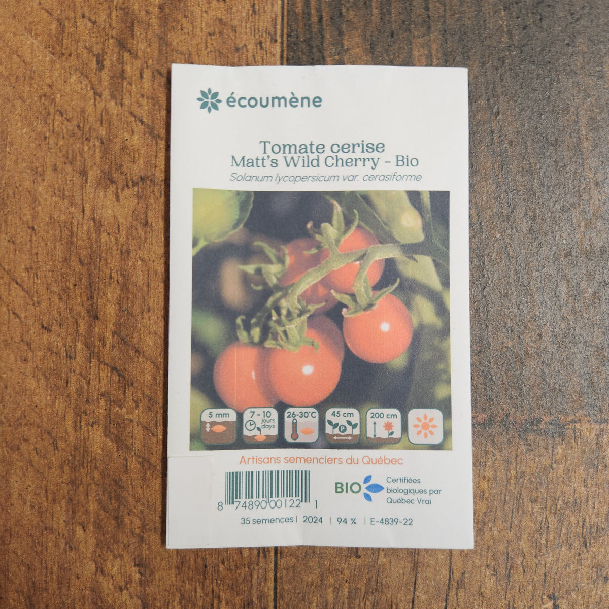Sachet de semences Tomate Cerise Matts Wild Cherry. Emballage blanc photographié de face sur un fond de bois brun. Une image du végétal occupe la partie centrale, sous le nom imprimé. De petits pictogrammes et un code-barres apparaissent au bas du sachet. La marque Écoumène figure dans la partie supérieure de cette présentation.