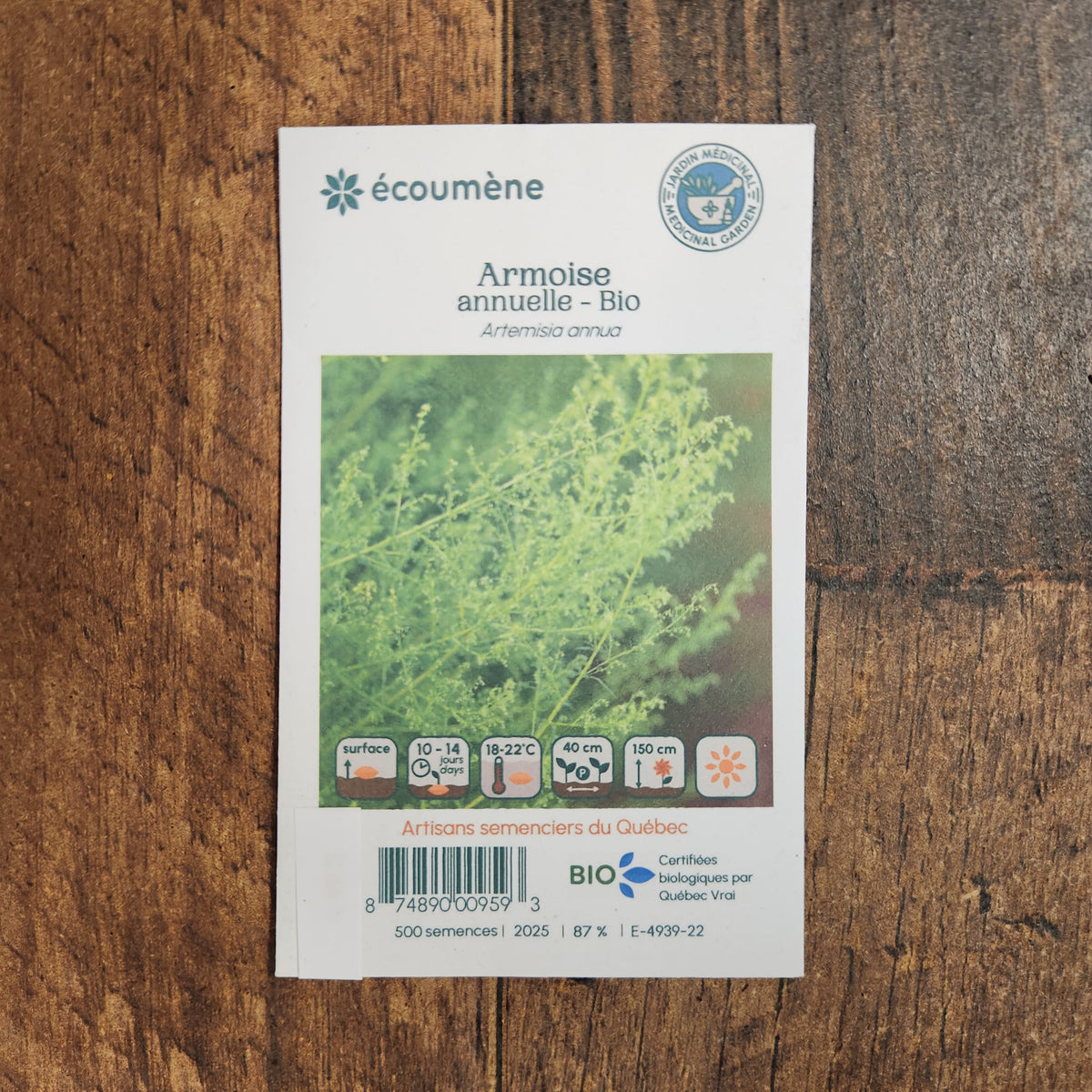 Sachet de semences Armoise annuelle. Emballage blanc photographié de face sur un fond de bois brun. Une image du végétal occupe la partie centrale, sous le nom imprimé. De petits pictogrammes et un code-barres apparaissent au bas du sachet. La marque Écoumène figure dans la partie supérieure de cette présentation.