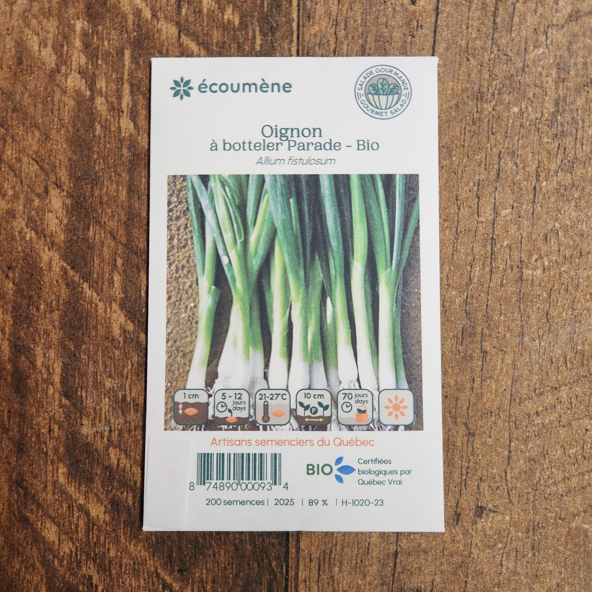Sachet de semences Oignon à botteler Parade. Emballage blanc photographié de face sur un fond de bois brun. Une image du végétal occupe la partie centrale, sous le nom imprimé. De petits pictogrammes et un code-barres apparaissent au bas du sachet. La marque Écoumène figure dans la partie supérieure de cette présentation.