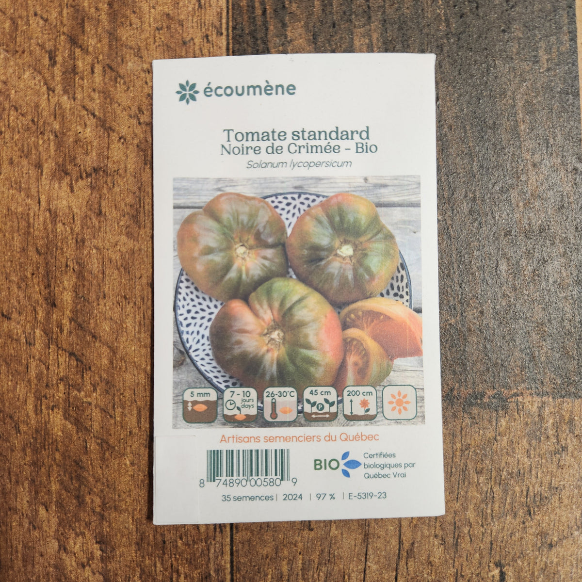 Sachet de semences Tomate Noire de Crimée. Emballage blanc photographié de face sur un fond de bois brun. Une image du végétal occupe la partie centrale, sous le nom imprimé. De petits pictogrammes et un code-barres apparaissent au bas du sachet. La marque Écoumène figure dans la partie supérieure de cette présentation.