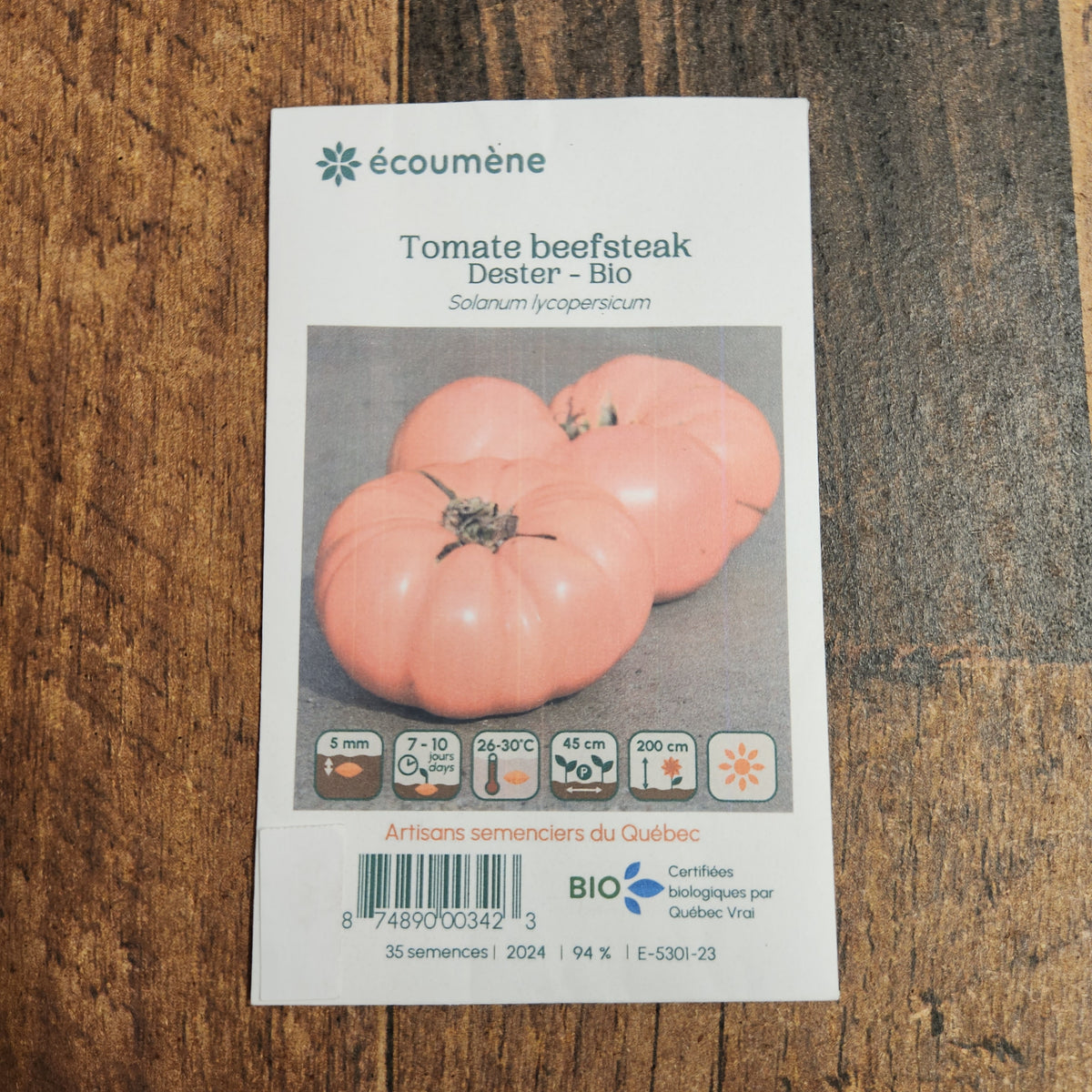 Sachet de semences Tomate Beefsteak Dester. Emballage blanc photographié de face sur un fond de bois brun. Une image du végétal occupe la partie centrale, sous le nom imprimé. De petits pictogrammes et un code-barres apparaissent au bas du sachet. La marque Écoumène figure dans la partie supérieure de cette présentation.