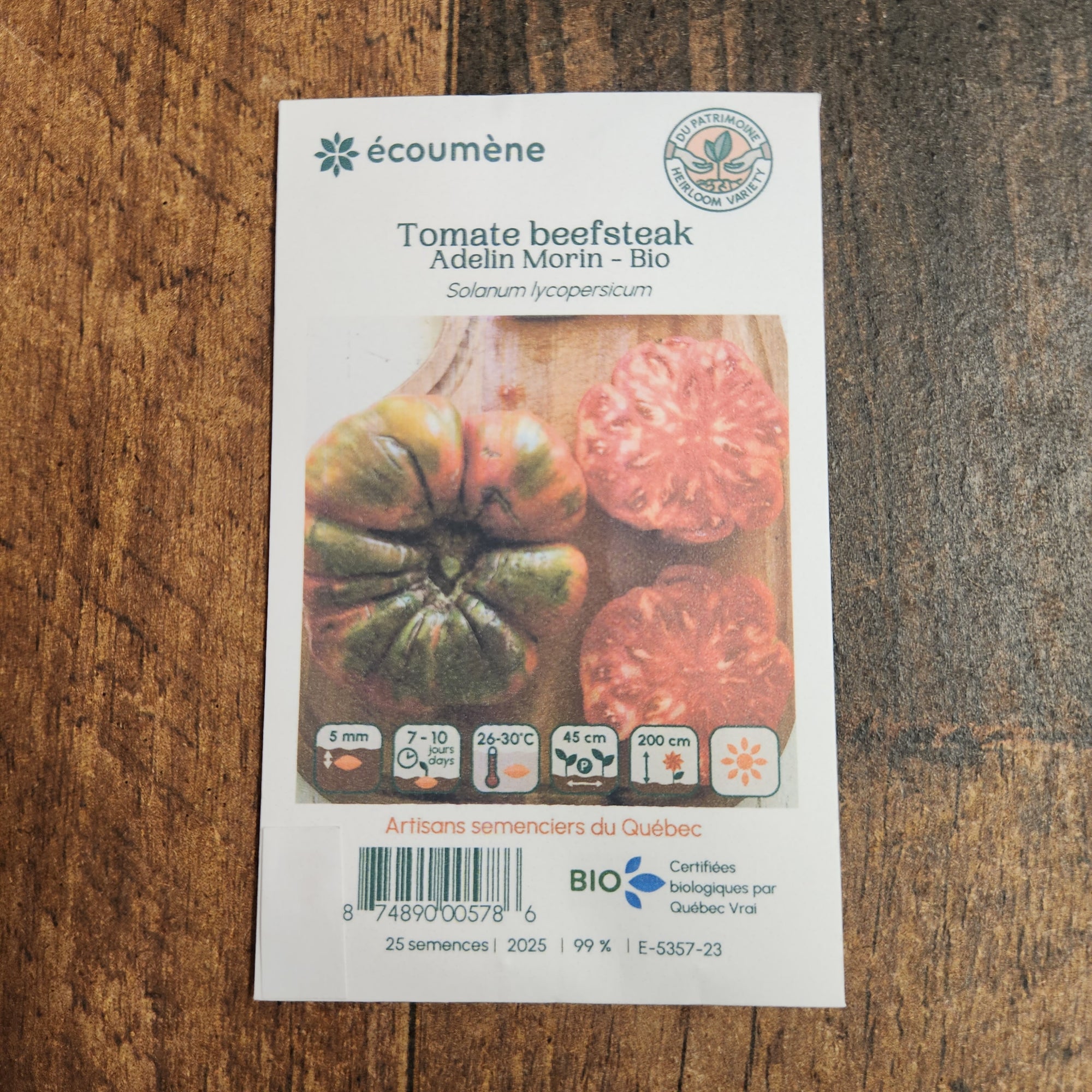 Sachet de semences Tomate Beefsteak Adelin Morin. Emballage blanc photographié de face sur un fond de bois brun. Une image du végétal occupe la partie centrale, sous le nom imprimé. De petits pictogrammes et un code-barres apparaissent au bas du sachet. La marque Écoumène figure dans la partie supérieure de cette présentation.
