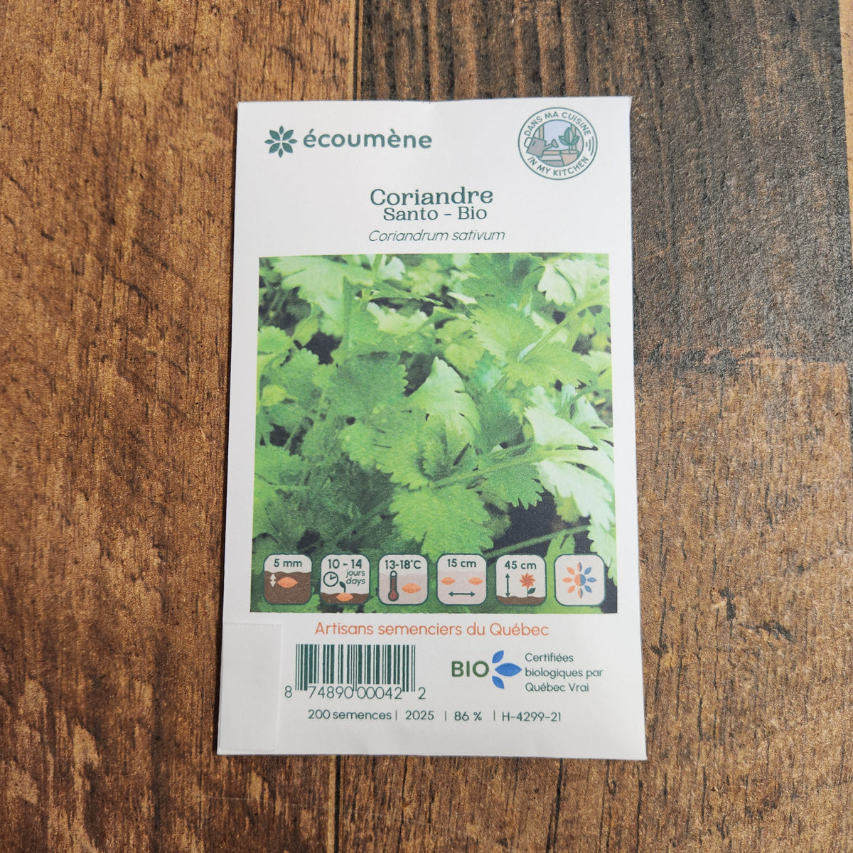 Sachet de semences Coriandre Santo. Emballage blanc photographié de face sur un fond de bois brun. Une image du végétal occupe la partie centrale, sous le nom imprimé. De petits pictogrammes et un code-barres apparaissent au bas du sachet. La marque Écoumène figure dans la partie supérieure de cette présentation.