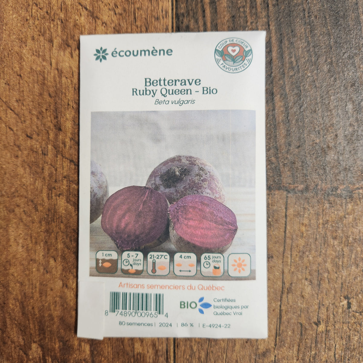 Sachet de semences Betterave Ruby Queen. Emballage blanc photographié de face sur un fond de bois brun. Une image du végétal occupe la partie centrale, sous le nom imprimé. De petits pictogrammes et un code-barres apparaissent au bas du sachet. La marque Écoumène figure dans la partie supérieure de cette présentation.