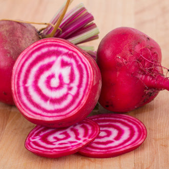 Whole and sliced organic 'Chioggia' beets showing pale red skin, concentric raspberry-pink and creamy-white rings, and edible green leaves with pink-striped stalks in a sunny harvest scene. The image highlights the slightly flattened round shape, smooth skin, regular rings, and delicate colour contrast that distinguish this heirloom variety when freshly cut for serving.