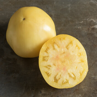 A whole pale-yellow Great White tomato sits behind a cut half on a dark, weathered surface. The whole fruit has smooth, softly rounded shoulders and a small dark mark near its top. The cross-section reveals cream-yellow flesh, a broad pale centre, and numerous seed chambers arranged around the outer portion.